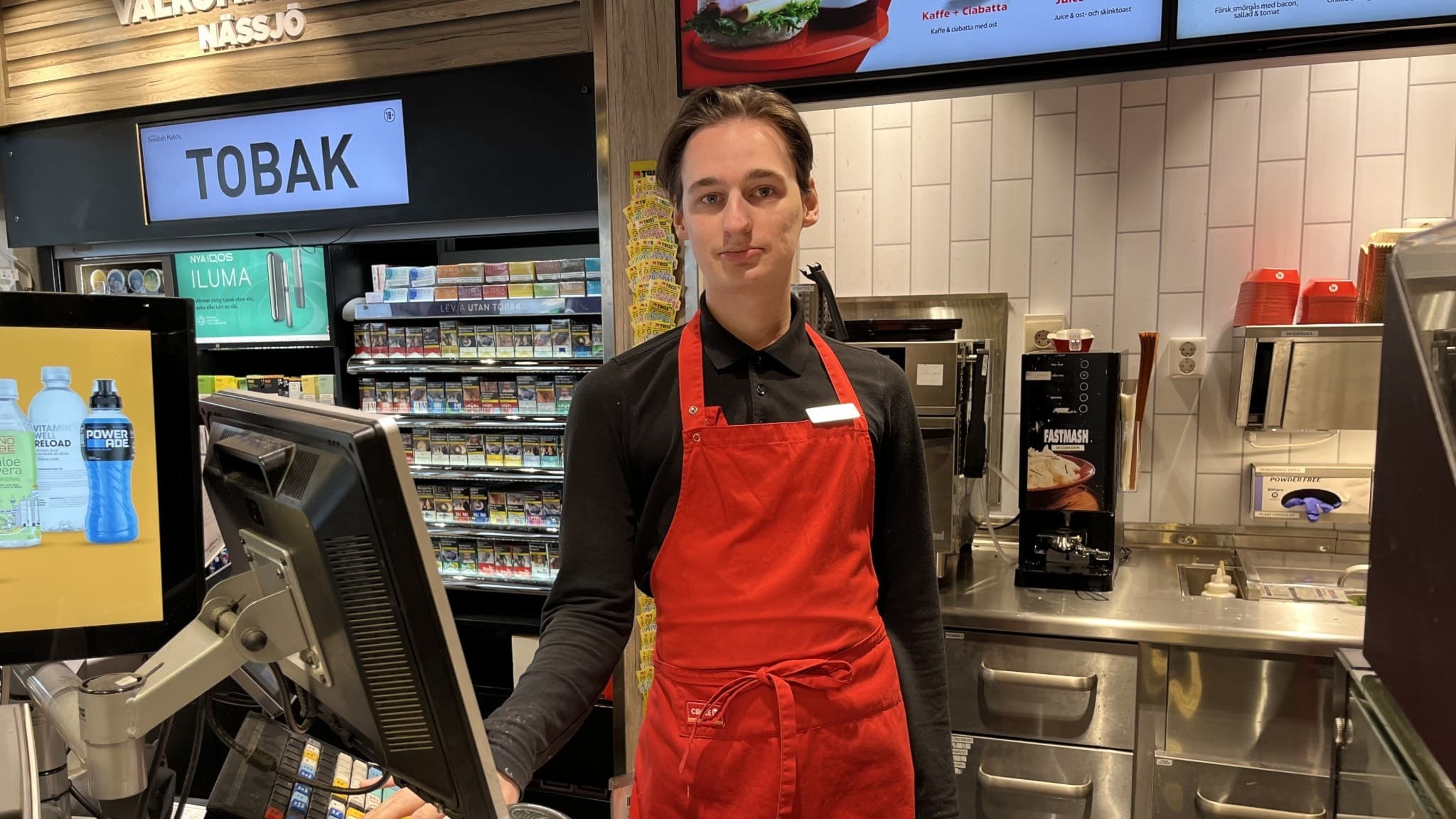 Edvin Olsson står med ett rött förkläde bakom kassan på macken.