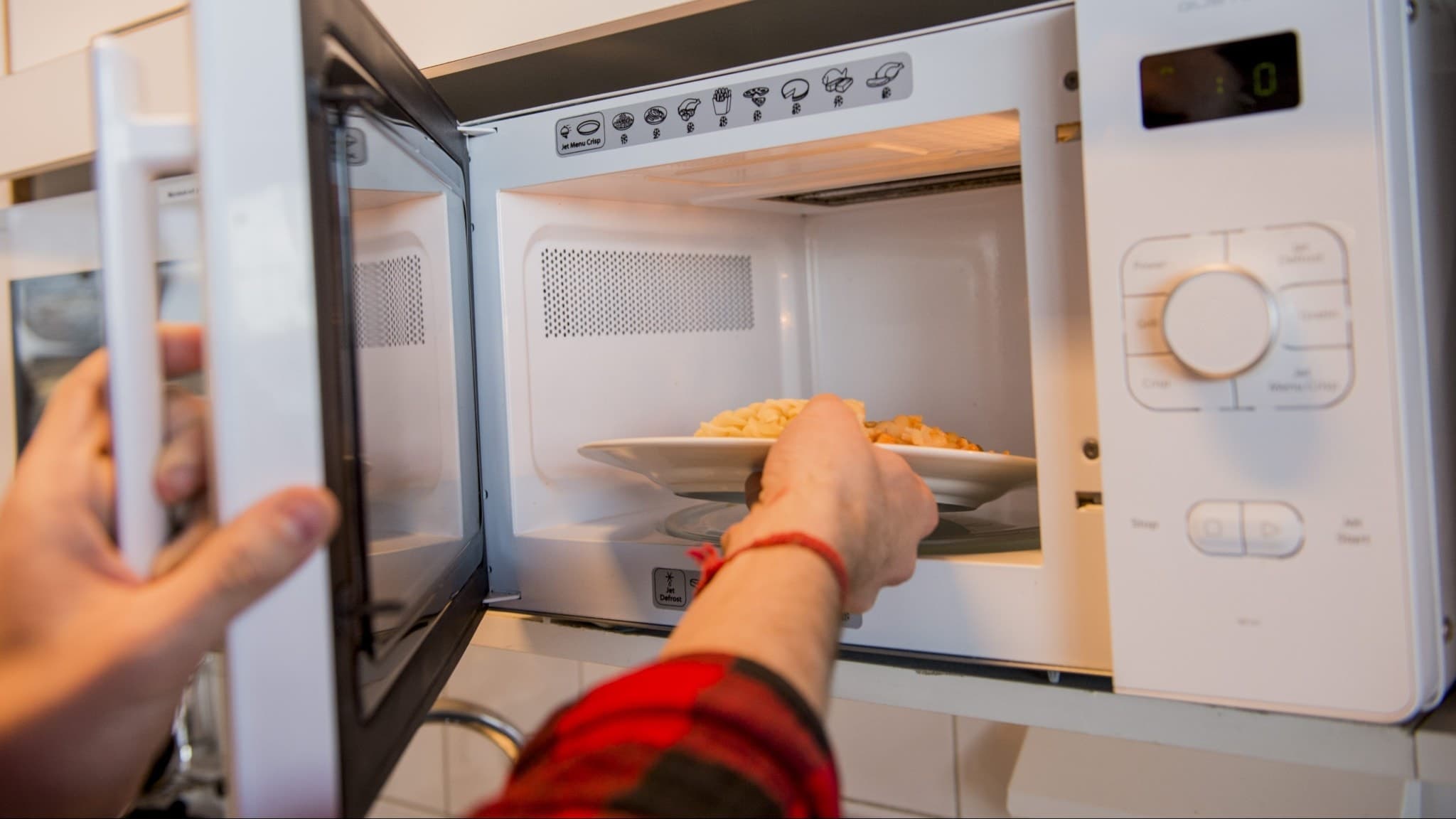En person i röd skjorta värmer en tallrik med mat i en mikrovågsugn på en arbetsplats. Mikrovågsugnen är vit med en öppnad lucka och synliga kontrollknappar.