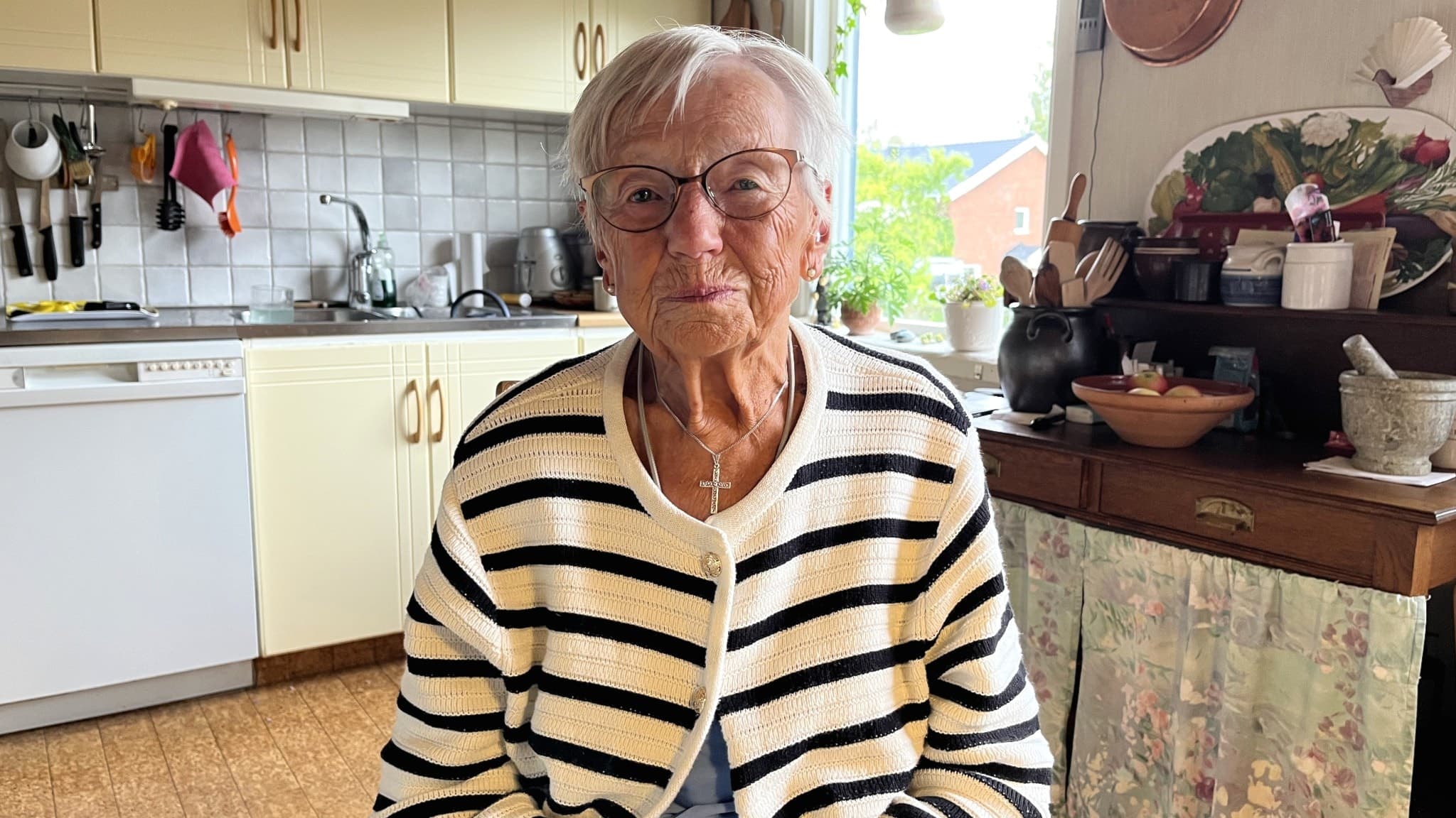 Mirjam Ring sitter i ett lantligt kök med ljusa skåp och en rutigt klädd köksbänk i bakgrunden. Hon bär en randig tröja och ett halsband.