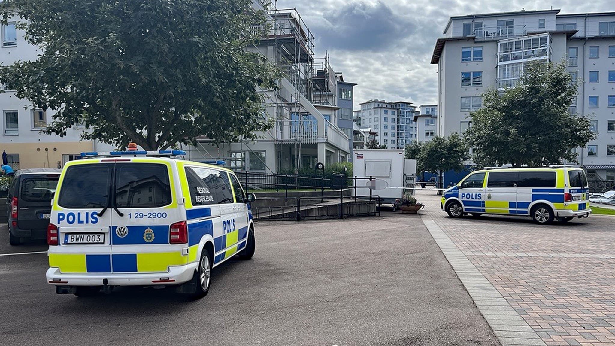 Två polisfordon står parkerade på ett torg i ett bostadsområde med flerfamiljshus i bakgrunden. En avspärrning är uppsatt, och några personer syns i närheten av platsen.