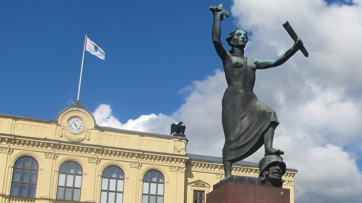 Fredsmonumentet i Karlstad på Stora torget, en kvinnofigur som håller ett avbrutet svärd. Hon står med vänsterfoten på ett huvud med soldathjälm. I bakgrunden syns en gul fasad med klocka och en flagga mot en blå himmel med vita moln.