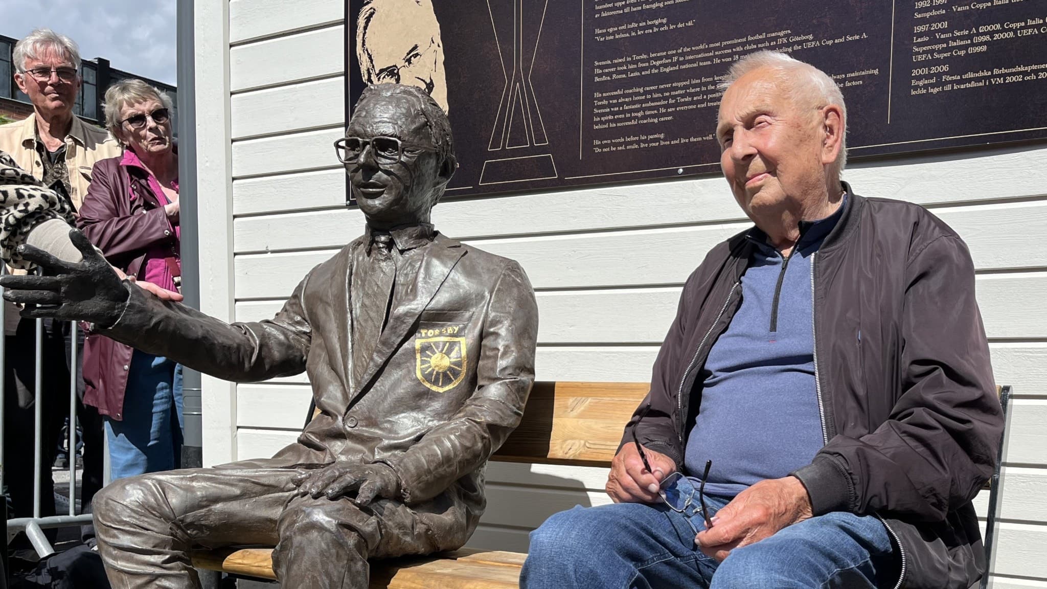 En äldre man sitter på en bänk bredvid en staty av Sven-Göran Eriksson, känd som Svennis. Statyn är klädd i kostym och gestikulerar med ena handen. Bakom syns en vägg med textskyltar.