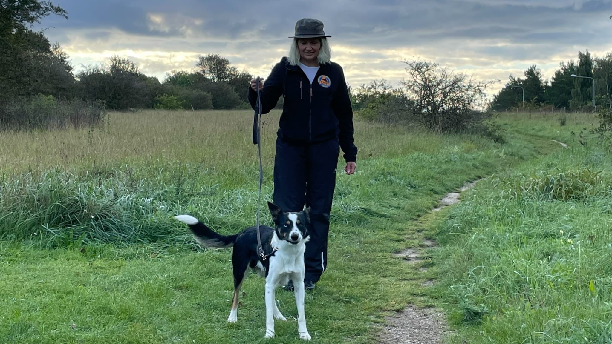 Anna-Karin Gustafsson promenerar med sin hund Tex på en grönskande stig i landsbygdsmiljö, under en molnig himmel. Hunden står alert bredvid henne med kopplet sträckt.