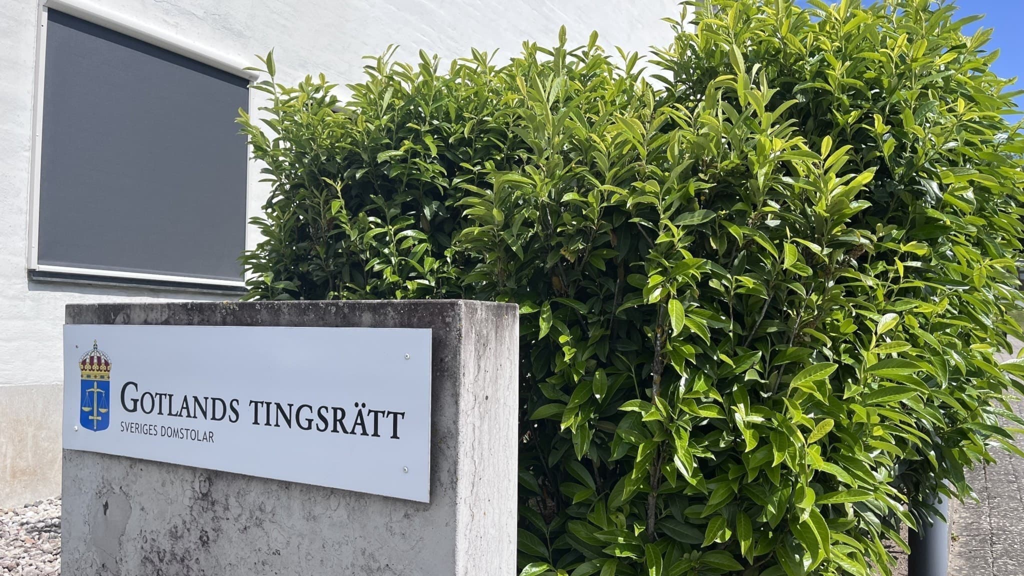 Byggnad med skylt som visar Gotlands tingsrätt, omgiven av grönskande buskar och en blå himmel i bakgrunden.