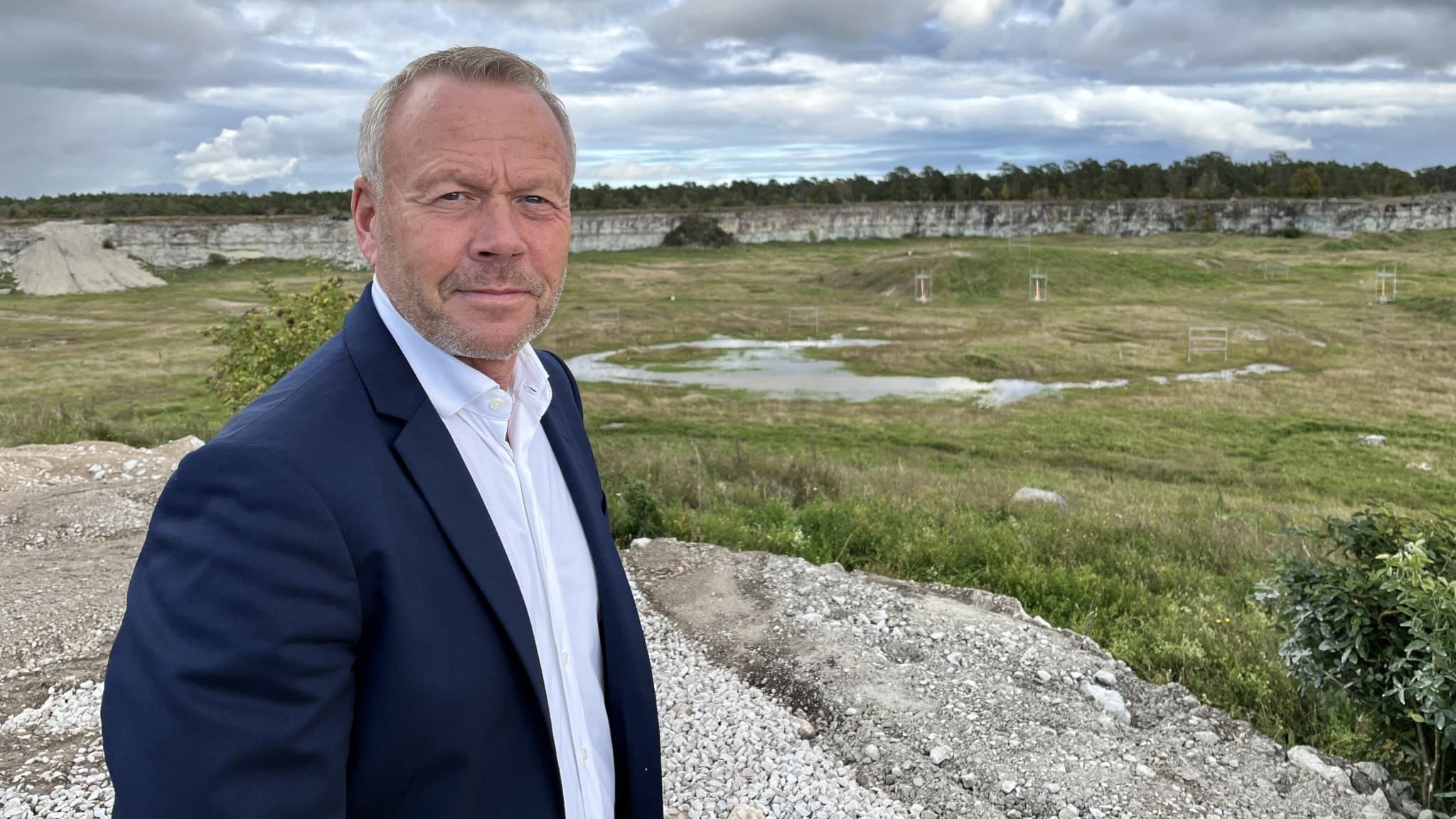 Conny Bohlin står vid den nya endurobanan i Hejdeby, ett grönskande kalkbrott med en dramatisk molnig himmel i bakgrunden, iklädd mörk kostym och vit skjorta.