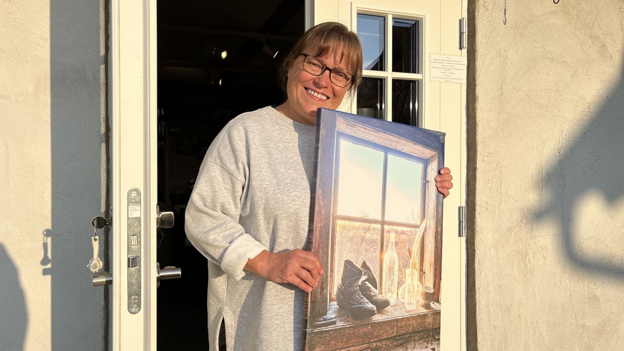 Ellen Marie står utanför en byggnad med dörren öppen och håller upp en tavla som föreställer en arkitektonisk scen med brovalv och natur. Solen lyser på henne och tavlan.