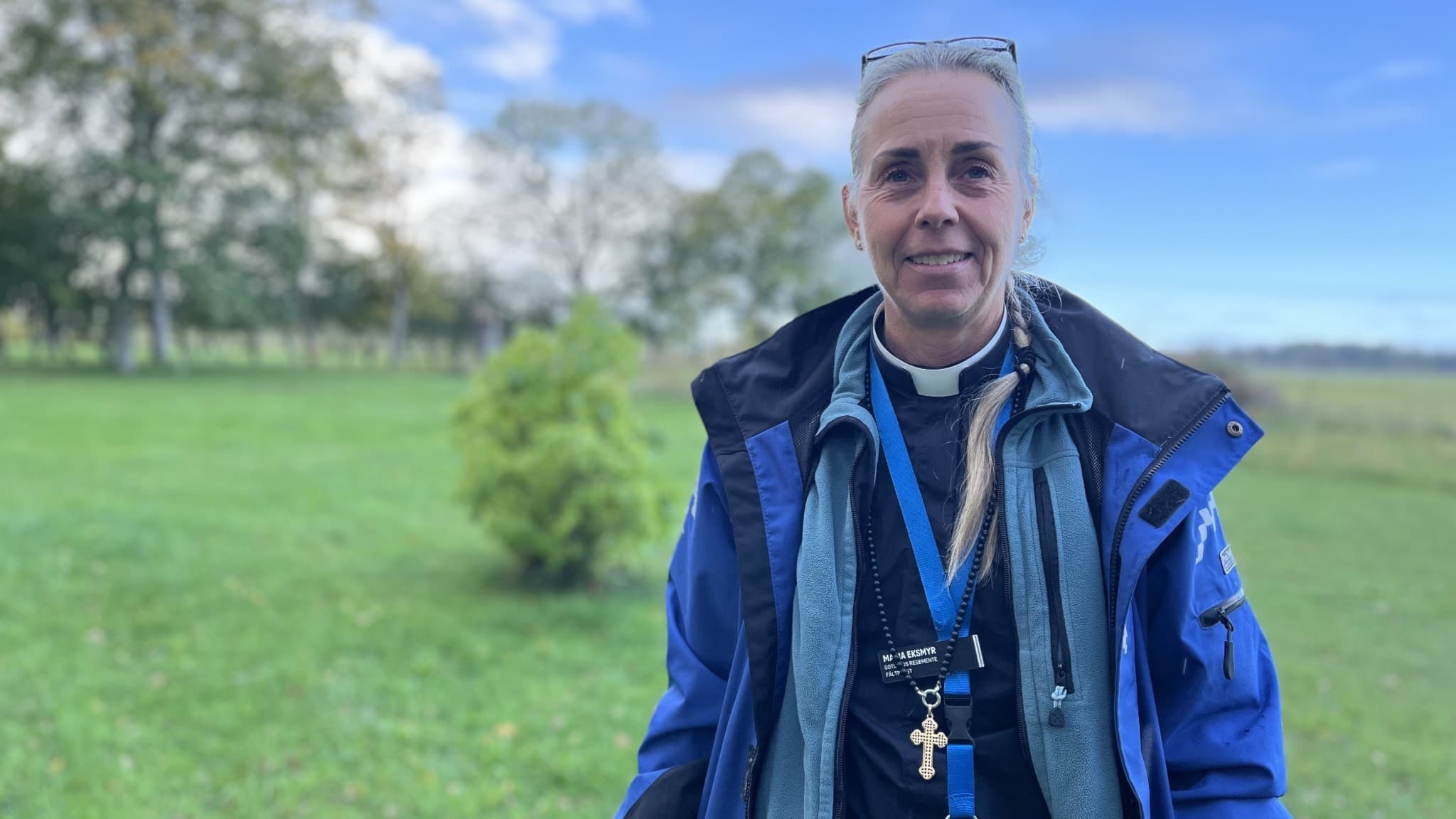 Maria Eksmyr står utomhus på en grön gräsmatta med träd och en blå himmel i bakgrunden. Hon har på sig en blå jacka, halsduk och ett blått nyckelband runt halsen.