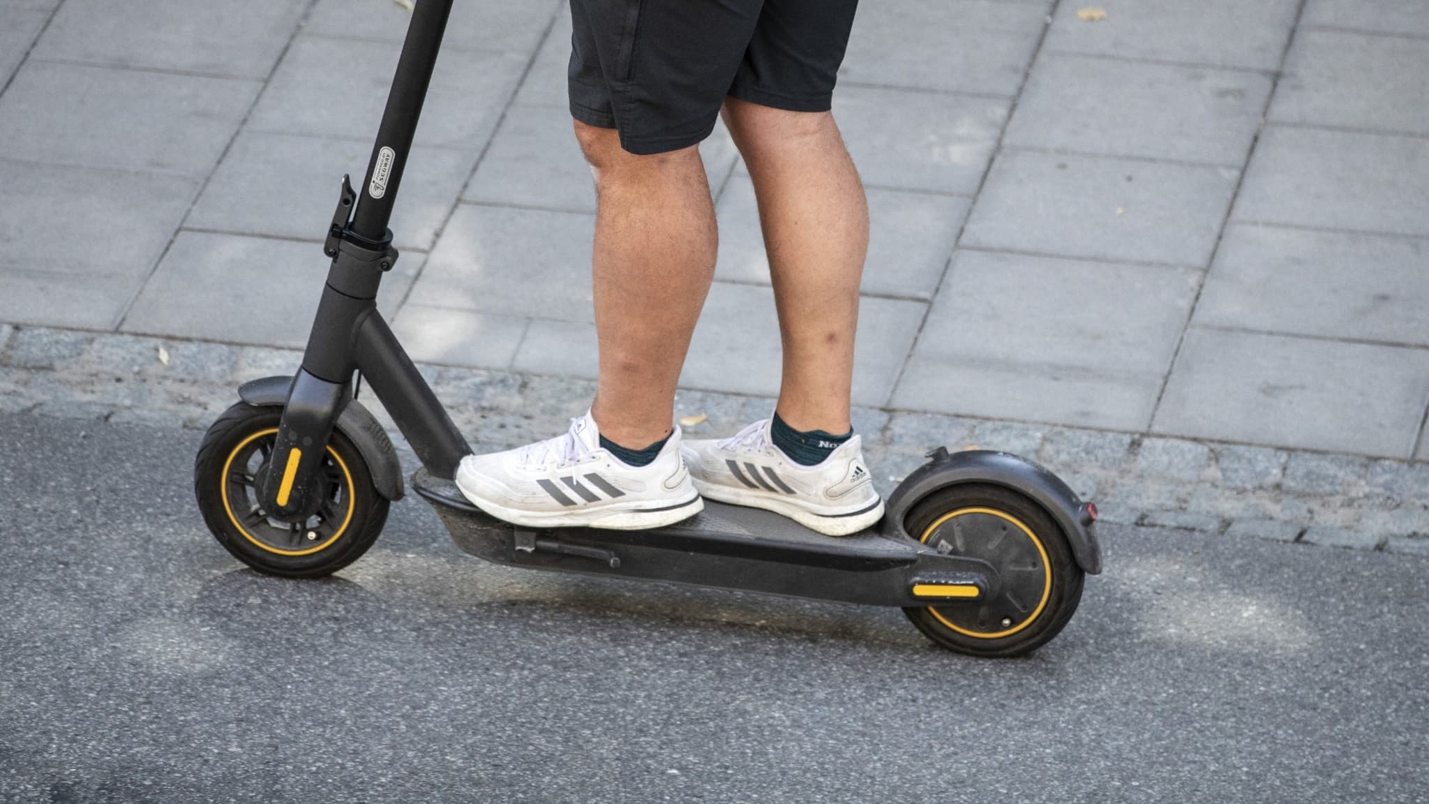 En person står på en elsparkcykel klädd i shorts och vita sneakers på en trottoar.