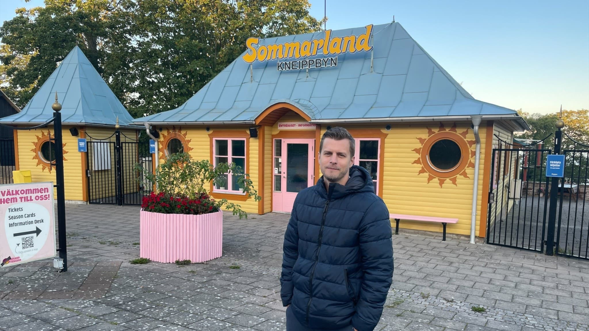 Simon Röcklinger står framför en gul byggnad med blått tak och solmotiv, som tillhör Summerhome. Miljön är utomhus med träd i bakgrunden och en stenlagd gårdsplan.