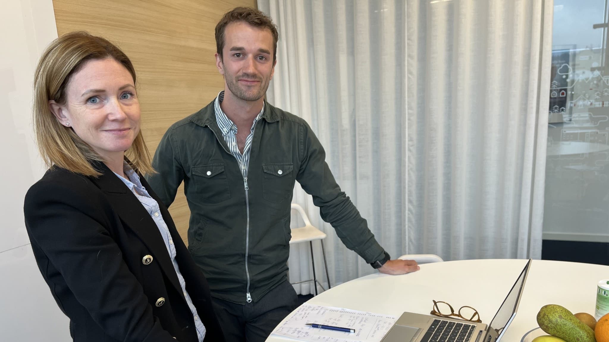 Malin Karlsson och Oskar Arnell, fastighetsmäklare på Länsförsäkringar Fastighetsförmedling, står vid ett bord med en laptop och fruktskål i ett modernt kontorsutrymme.