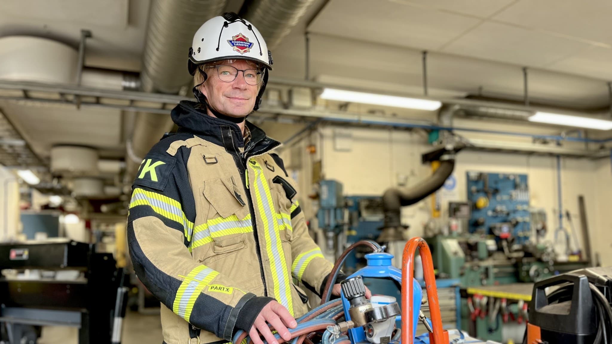 Brandmannen Per Tylemo, klädd i skyddsutrustning och hjälm, håller händerna på en gasflaska, i en verkstadsmiljö med maskiner och verktyg i bakgrunden.
