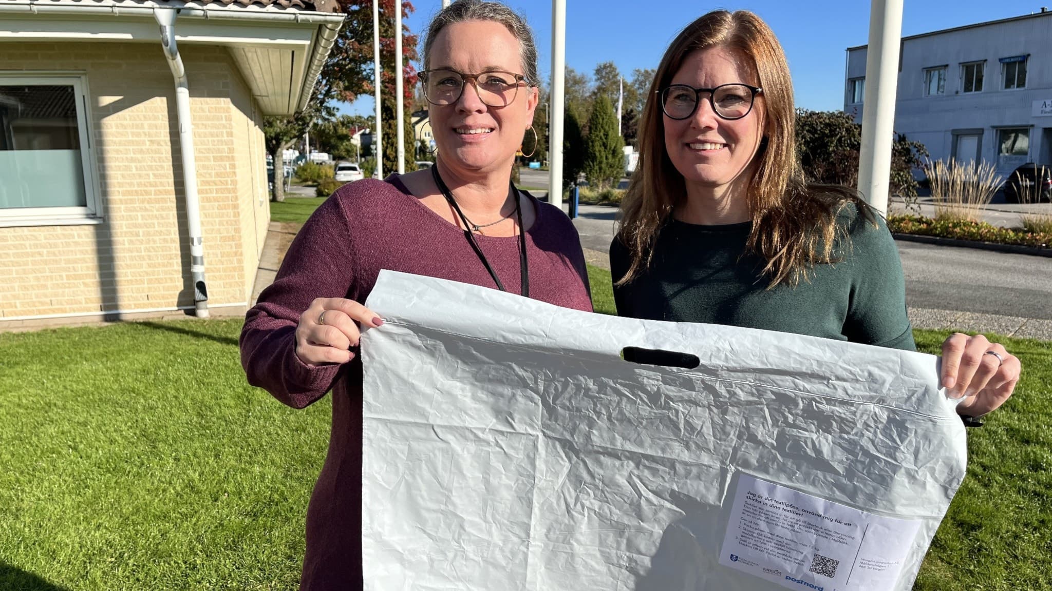 Sandra Brorsson från Mjöbäck visar en vit återvinningspåse för textilier tillsammans med miljöstrategen Therese Silow. De står utomhus framför byggnader i soligt väder.
