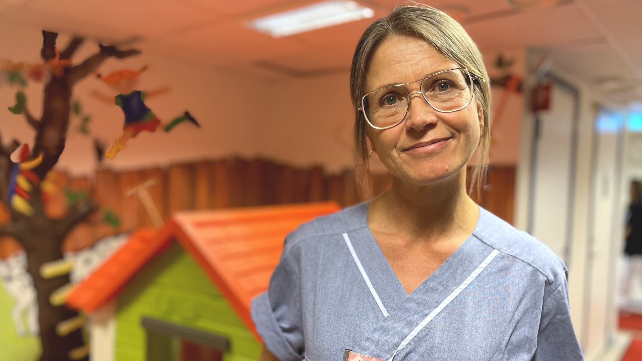 Camilla Damm, BHV-sjuksköterska på Närhälsans BVC, står i en färgglad miljö med barnmotiv och ett lekhus i bakgrunden, iklädd arbetskläder och namnskylt.