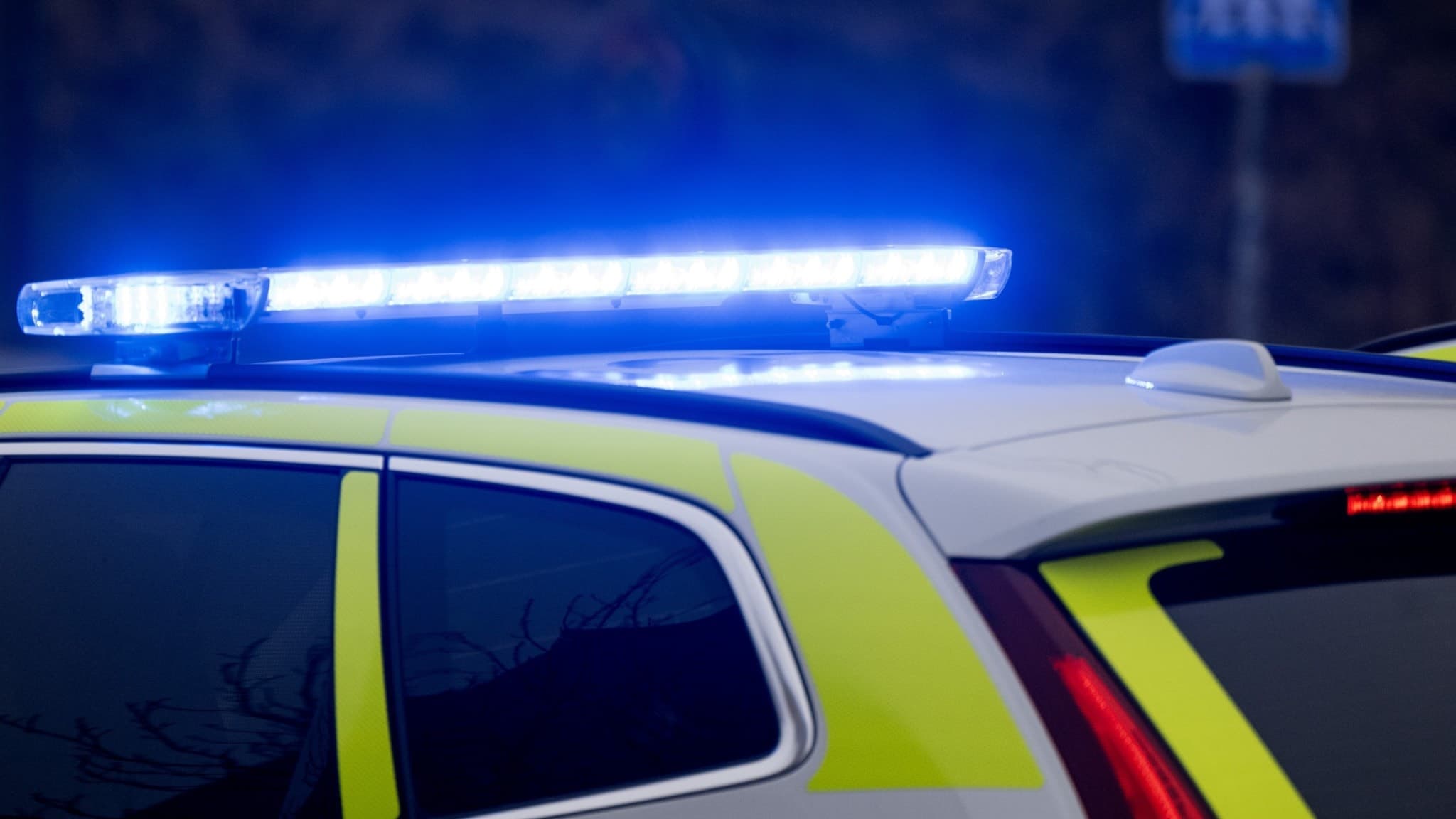 En polisbil med aktiverade blåljus står stilla. Fordonet har gul-blå markeringar och befinner sig vid en vägskylt. Bilden förmedlar en känsla av pågående polisinsats i urban miljö.