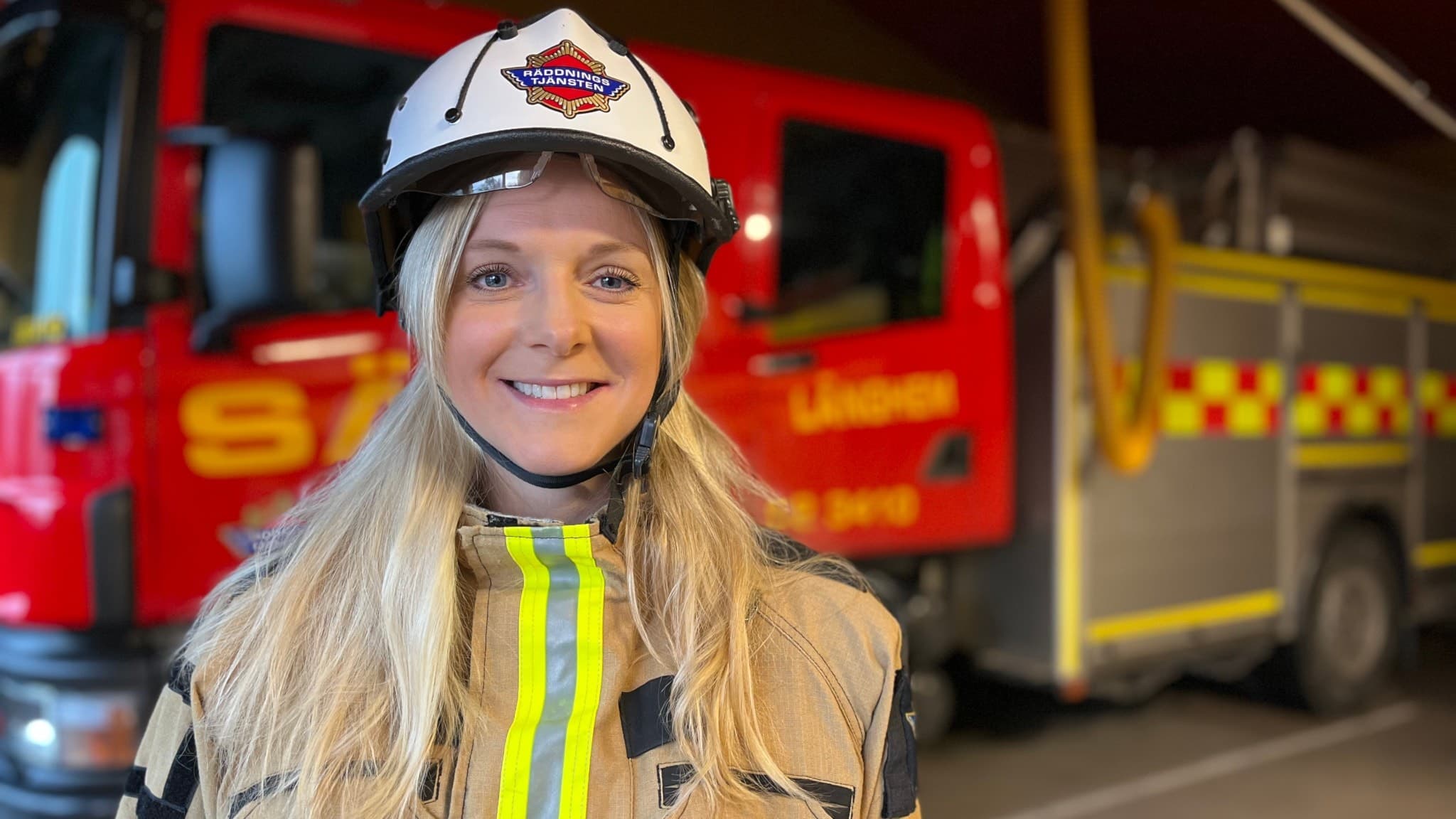 En brandkvinna i uniform och hjälm står framför en röd brandbil. Bilden visar en insatsmiljö kopplad till räddningstjänsten.