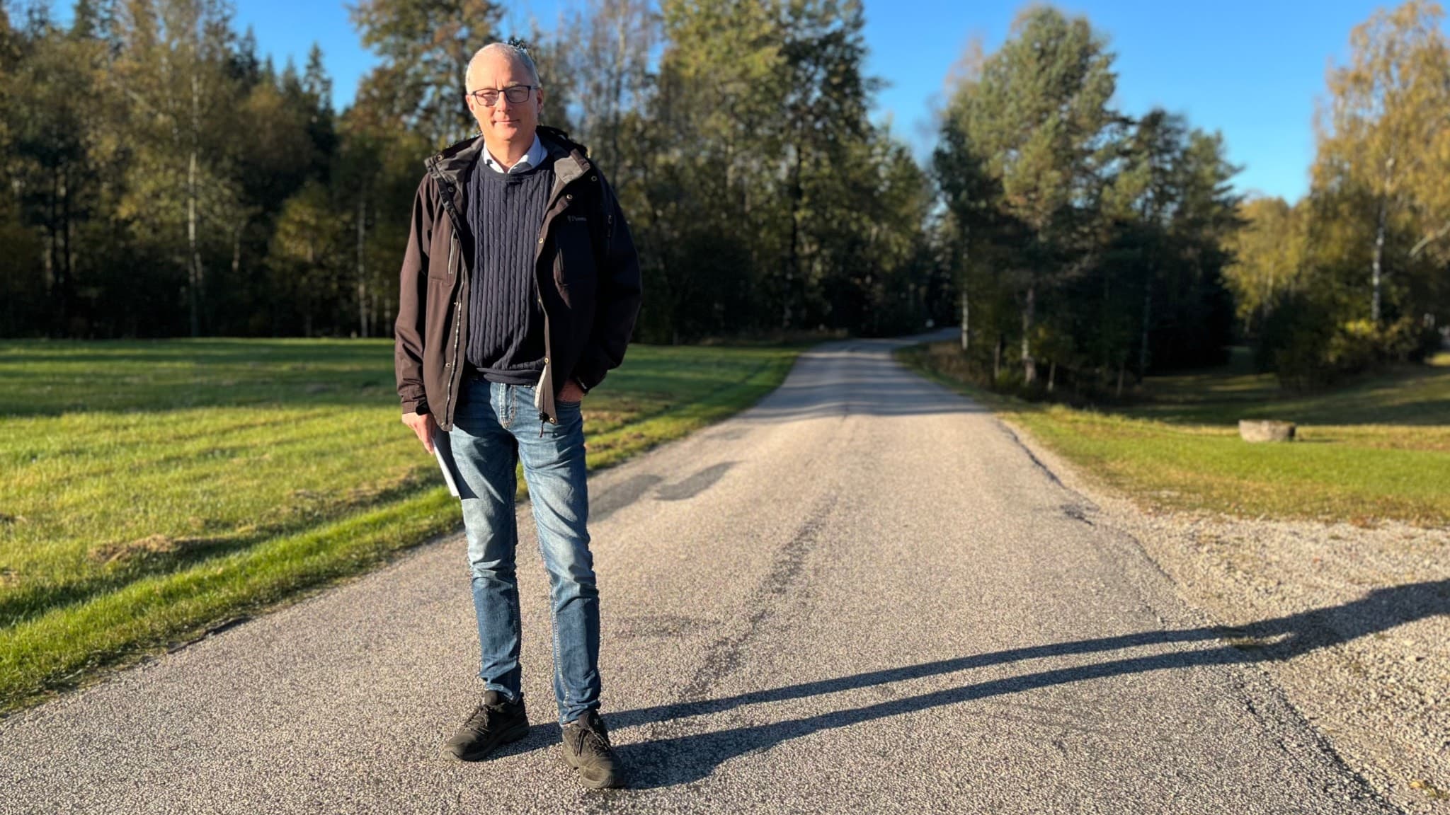 Stefan Magnusson, kassör i Hedared Vemmenhults vägsamfällighet, står på en enskild väg som ska byggas om. Solen skiner och omgivningen består av grönska och träd i bakgrunden.
