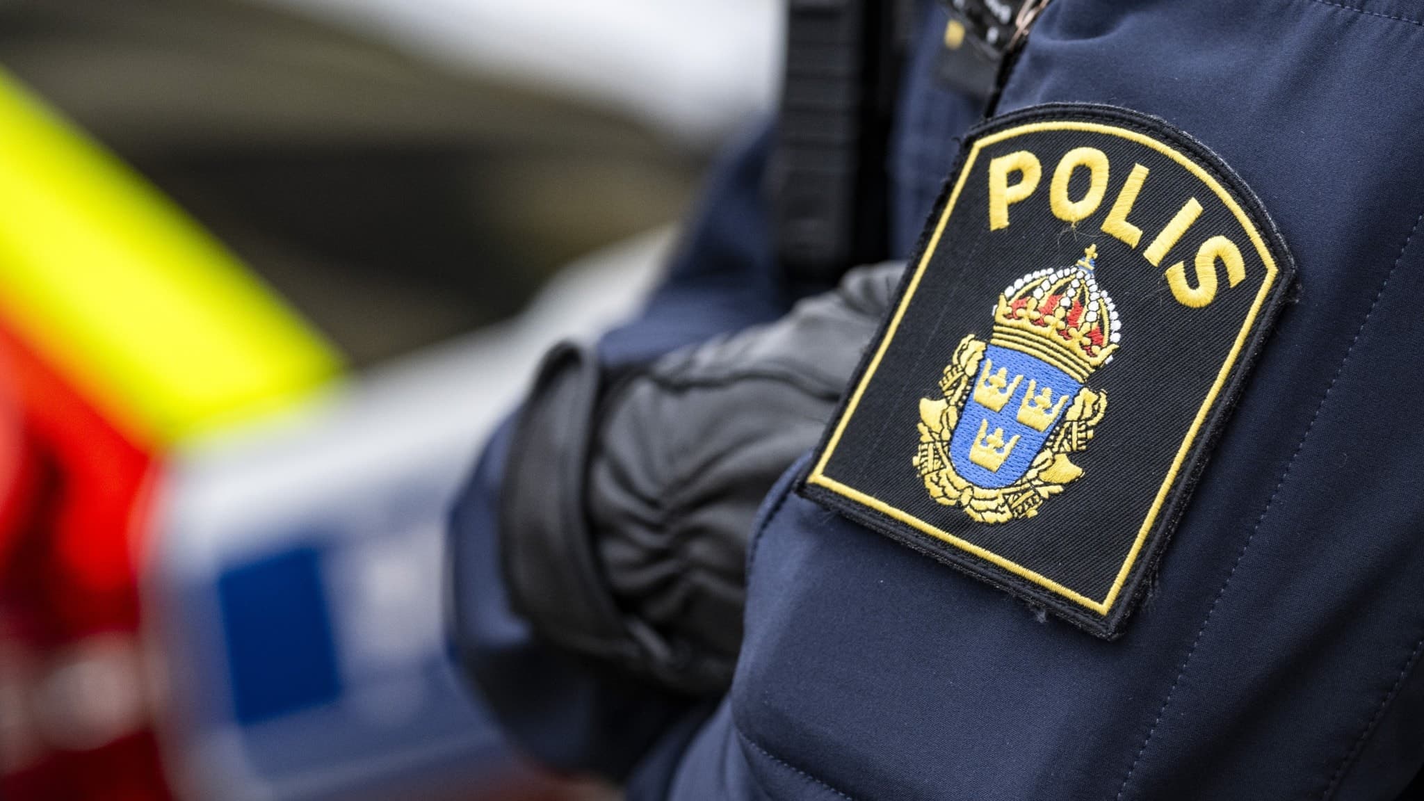 En detaljbild på en polisuniform med svensk polislogotyp synlig. I bakgrunden skymtar ett utryckningsfordon.