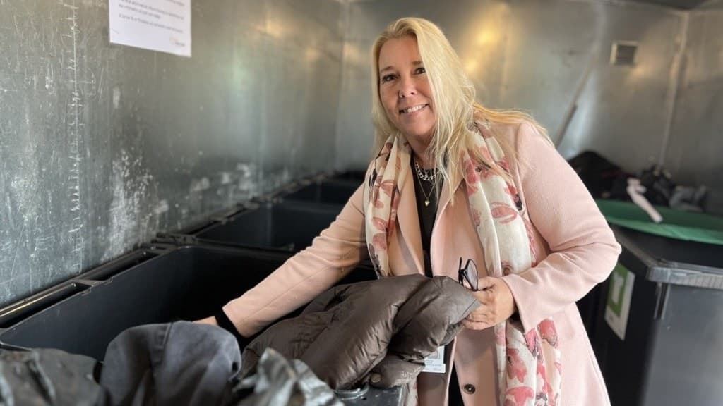 Jessica Petroff ses lägga en jacka i en återvinningsbehållare. Hon bär en ljusrosa kappa och står i en insamlingsstation för kläder, med flera behållare omkring sig.