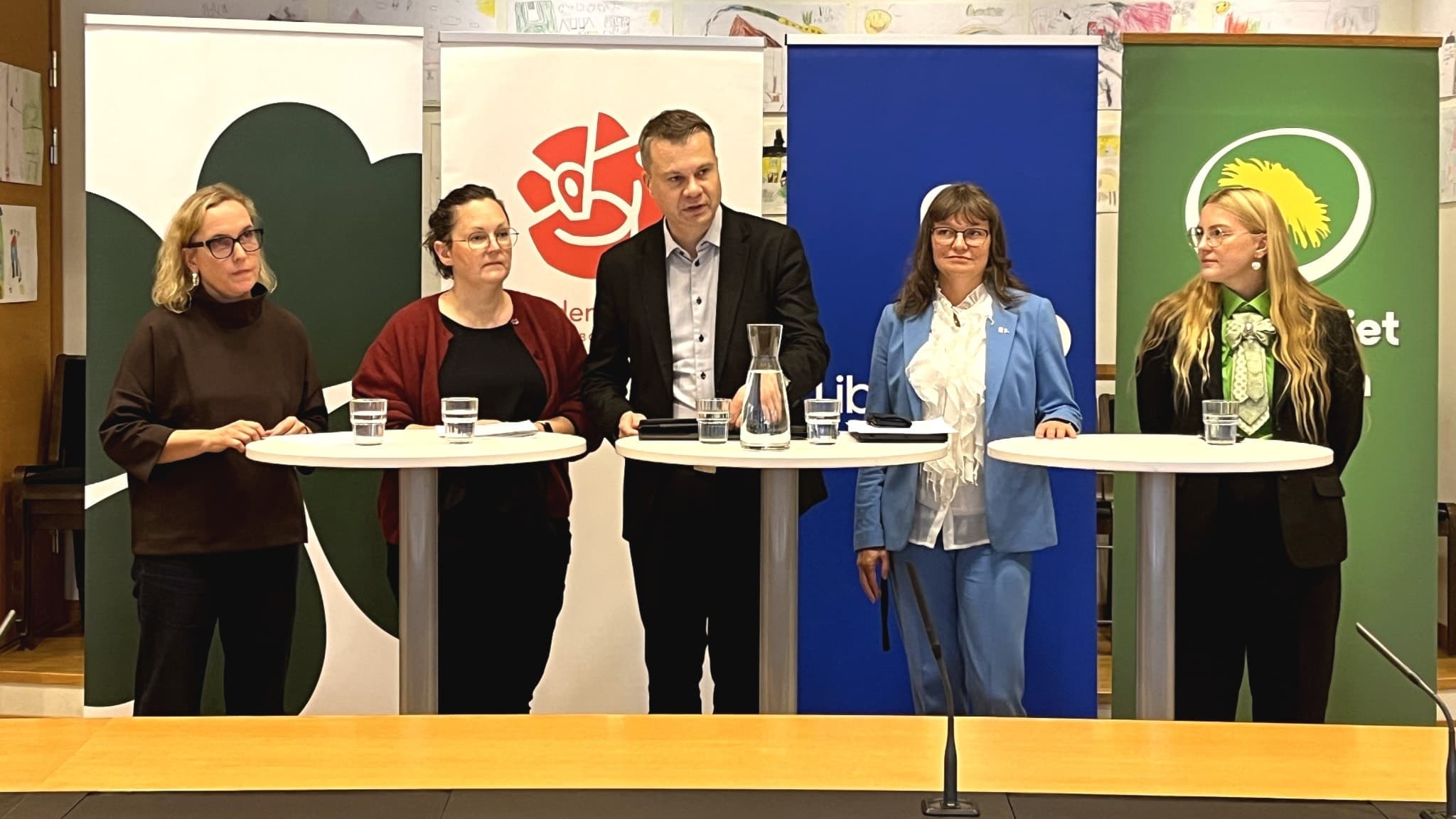 Fem politiker står vid podium med partiflaggor i bakgrunden, representerande Centerparitet, Socialdemokraterna, Liberalerna och Miljöpartiet under en presskonferens i Stadshuset.