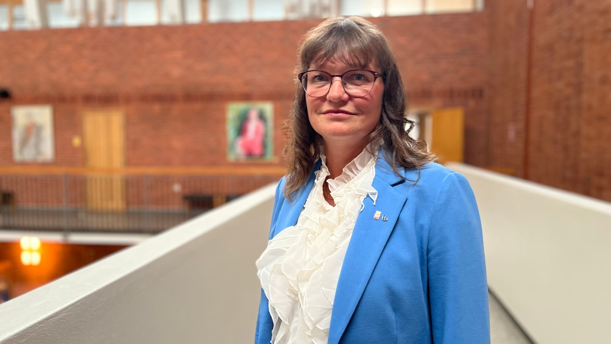 Anna Svalander står i en modern byggnad med tegelväggar. Hon är klädd i en blå kavaj och vit blus. I bakgrunden syns konstverk och glaspartier som släpper in naturligt ljus.