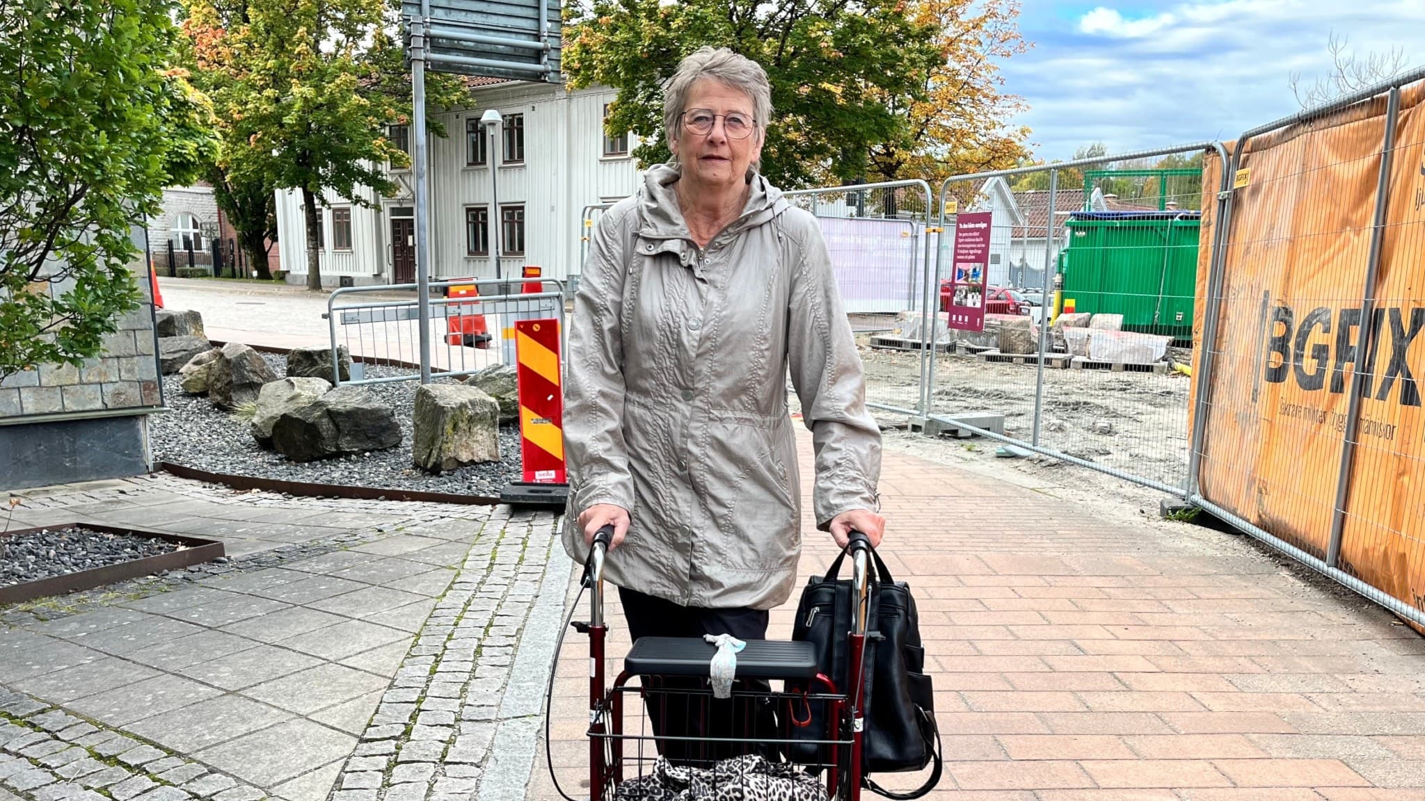 Britt Inger Eriksson går med en rullator på en trottoar intill ett pågående vägarbete med avspärrningar och byggstaket i bakgrunden.