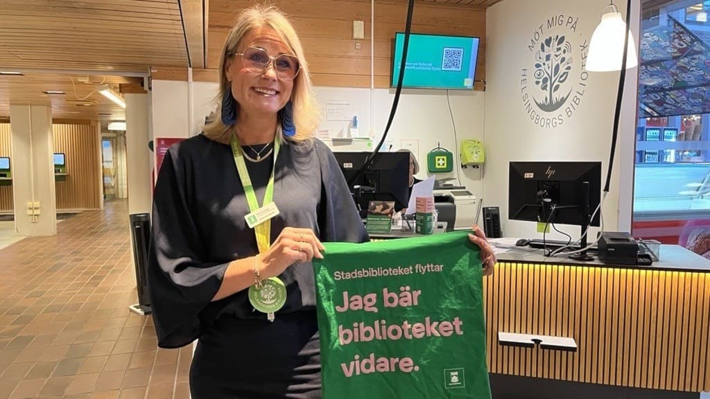 En person står i ett bibliotek och håller upp en grön tygpåse med texten 'Jag bär biblioteket vidare' framför en receptionsdisk. Skylten bakom visar bibliotekets logotyp.