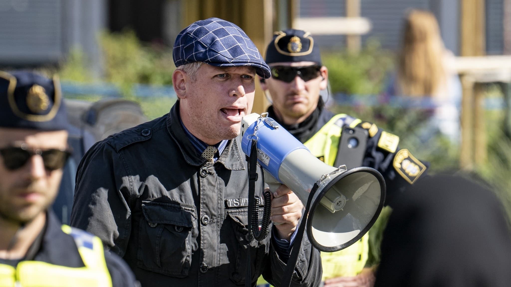 Rasmus Paludan står utomhus och pratar i megafon som han håller i handen. Flera poliser syns runtomkring.
