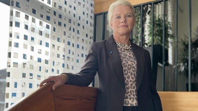 En person i kostym står inomhus vid ett trappräcke. Bakgrunden visar stora fönster och en modern byggnad utanför.