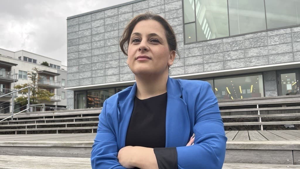 Tina Falk står i en blå kavaj står med armarna i kors framför en modern byggnad med stora glaspartier. I bakgrunden syns trappsteg av trä och en molnig himmel.