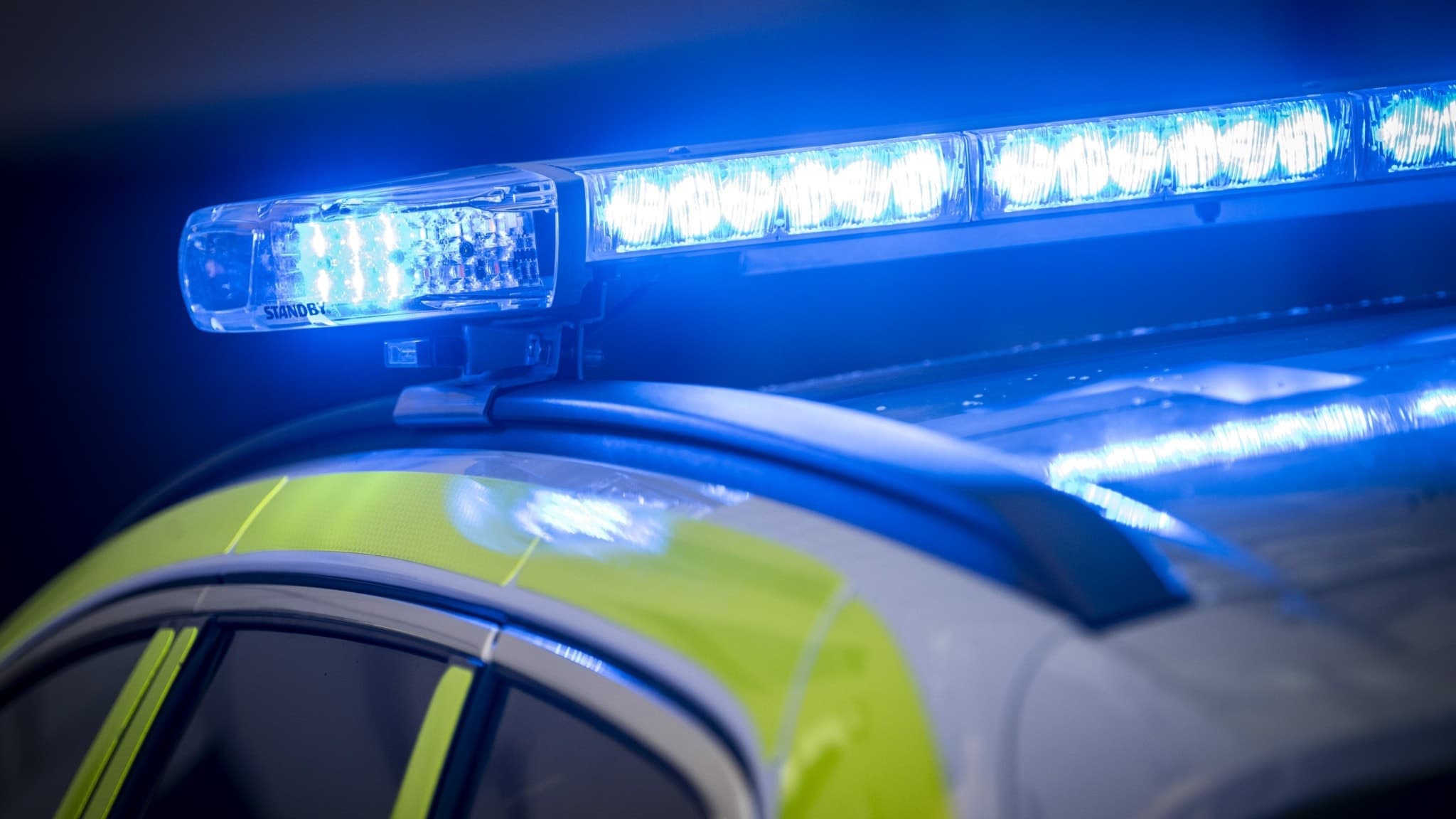Närbild på en polisbil med blåljus aktiverade, vilket lyser starkt i mörk omgivning. Fordonets gulgröna reflexdetaljer syns tydligt på taket.