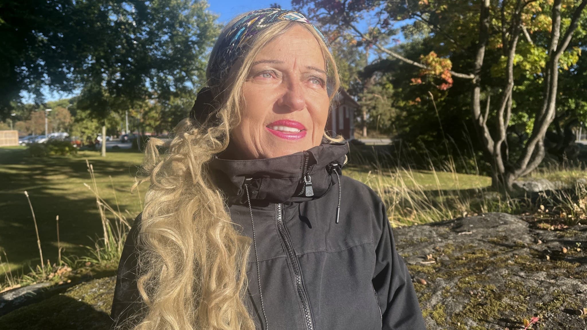 En person med långt, blont hår står utomhus i en park med färgglada höstträd i bakgrunden. Personen bär en svart jacka och det är soligt väder.