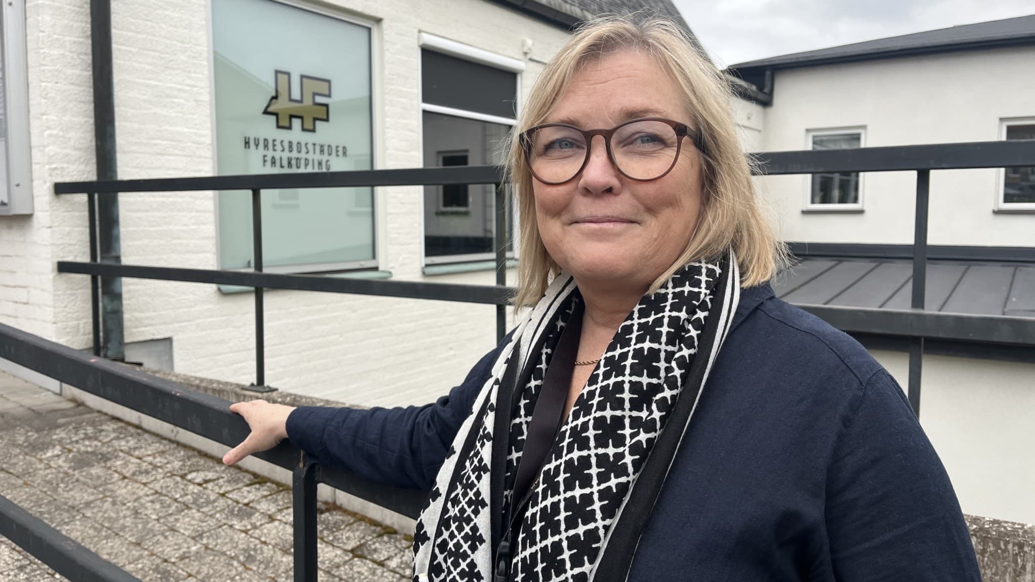 En person står framför en byggnad med en skylt där det står 'Försäkringskassan'. Personen är klädd i en mörk jacka och en svartvit halsduk. Byggnaden har vita väggar och svarta detaljer.