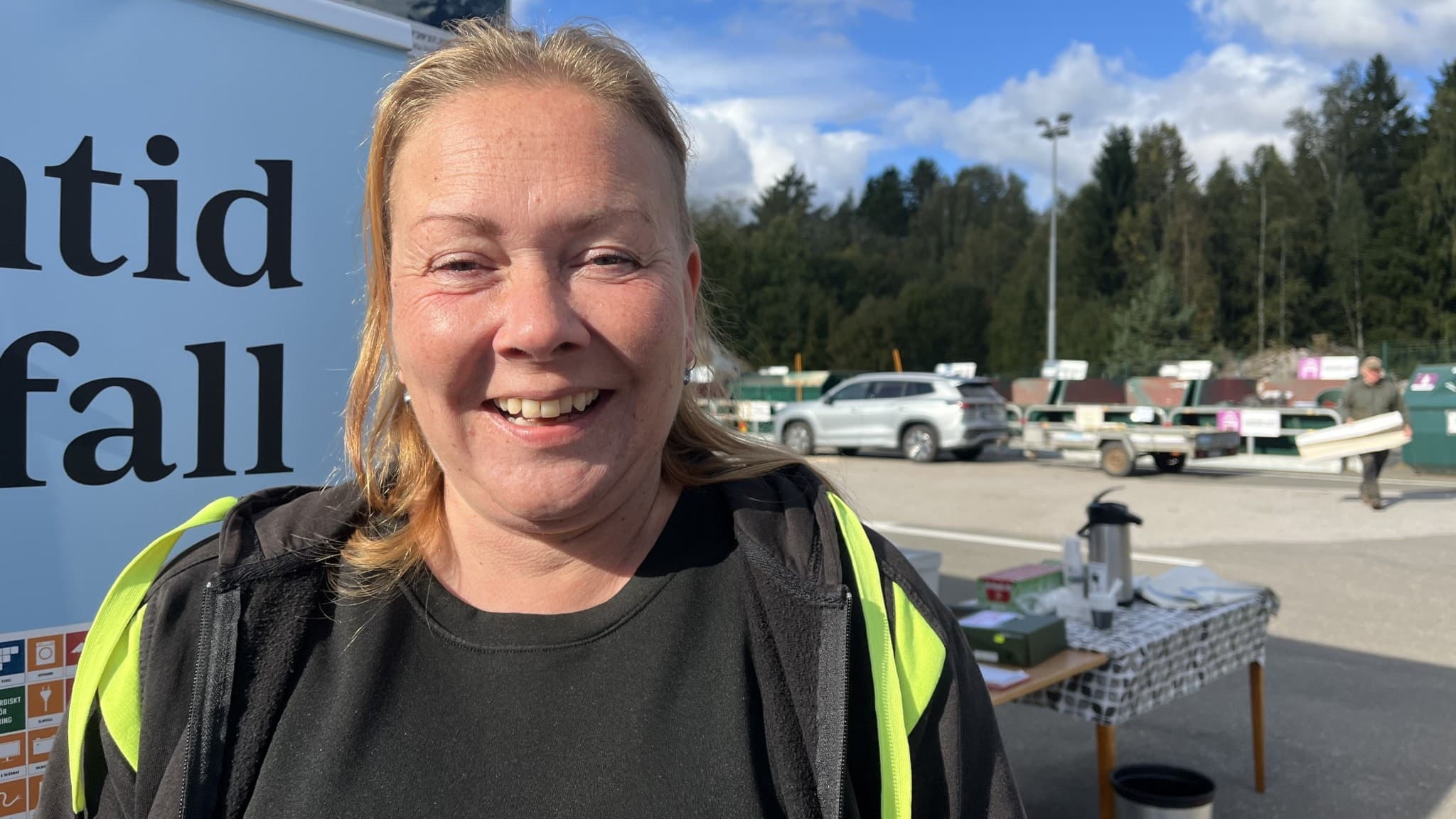 En kvinna med reflexväst står framför en skylt som delvis visar texten 'tid' och 'fall'. I bakgrunden syns en återvinningsstation med containrar, bord och skogsområde.