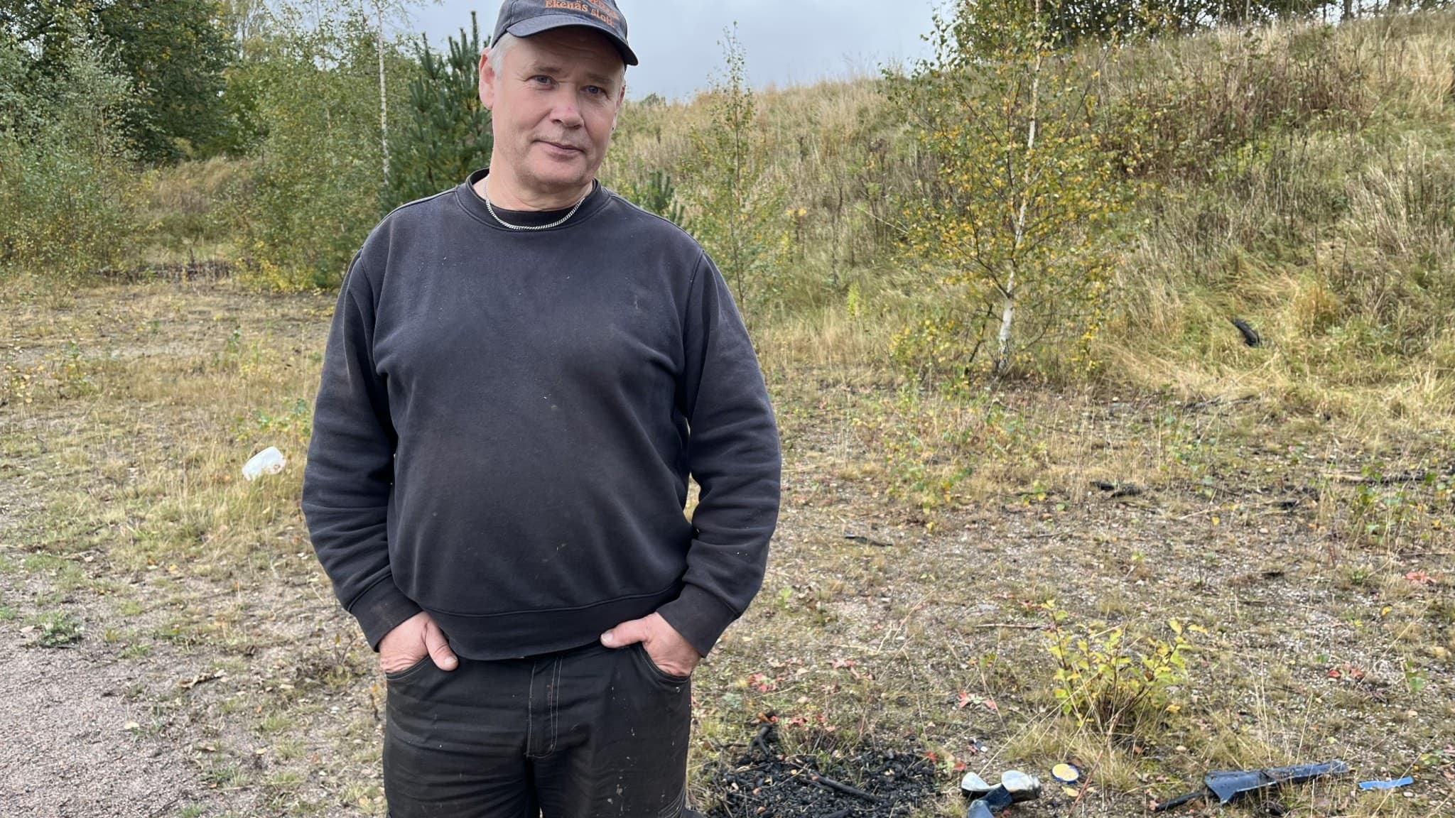 Roger Hellner står utomhus på en grusväg omgiven av gräs och buskar. Han är klädd i mörka kläder och har händerna i fickorna. Marken visar spår av nedskräpning och brännmärken.