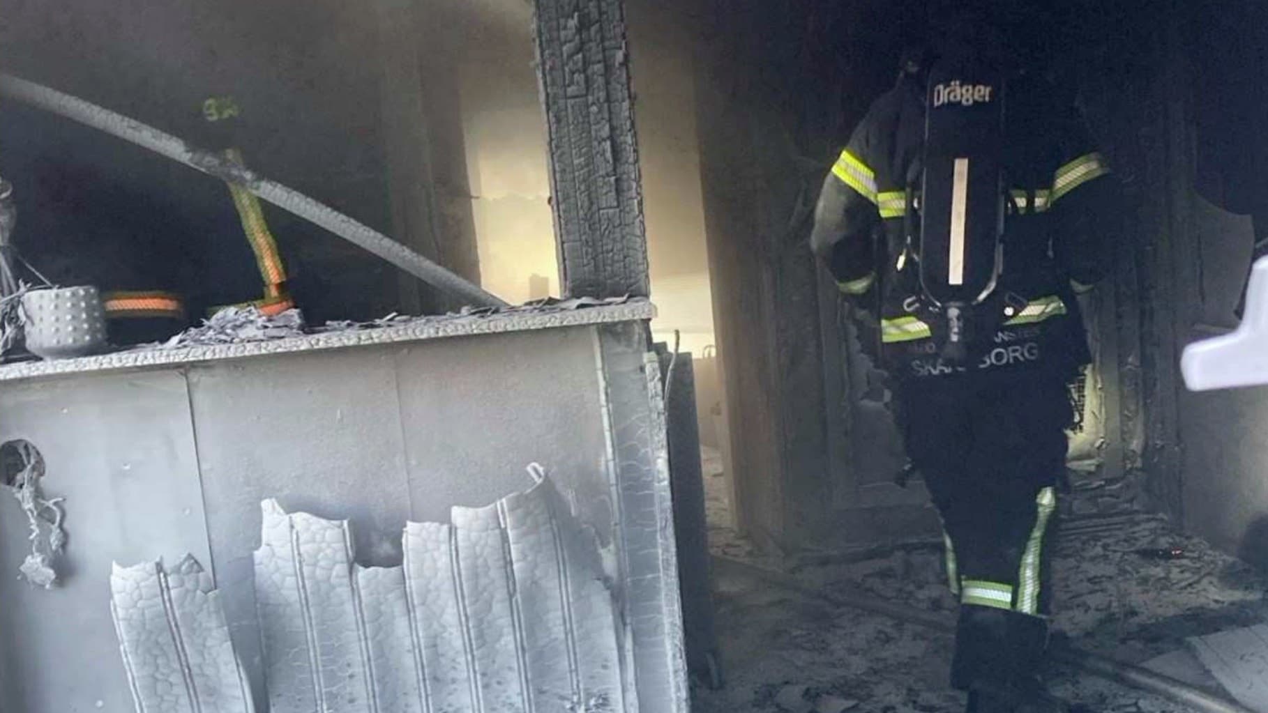En brandman i full utrustning rör sig genom ett brandskadat rum med sotiga väggar och förstörda möbler.