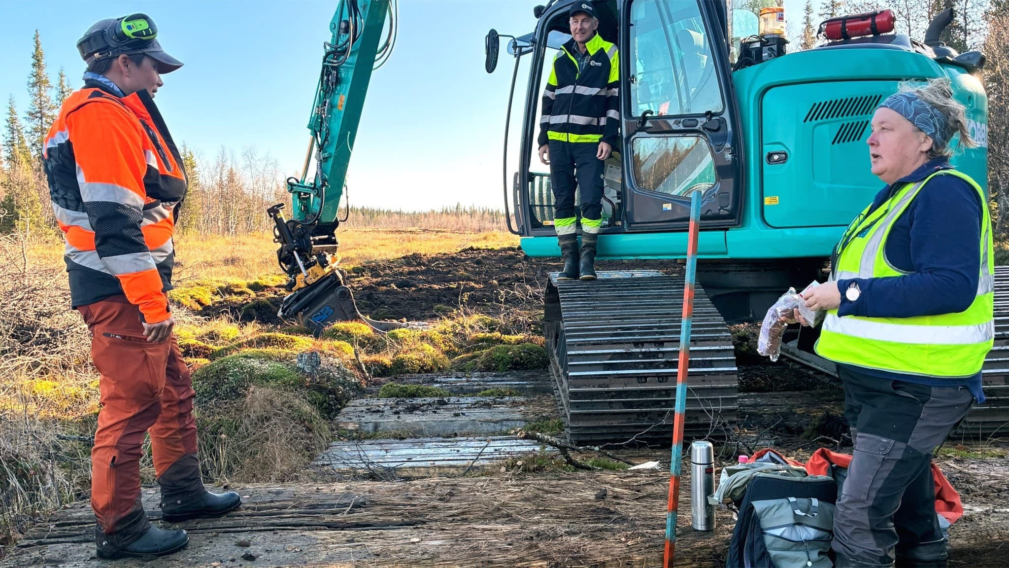 Tre personer i arbetskläder vid en grävmaskin som används för att återskapa en myr utanför Tjautjas i Gällivare kommun. Miljövård och återställning av våtmarker i fokus.