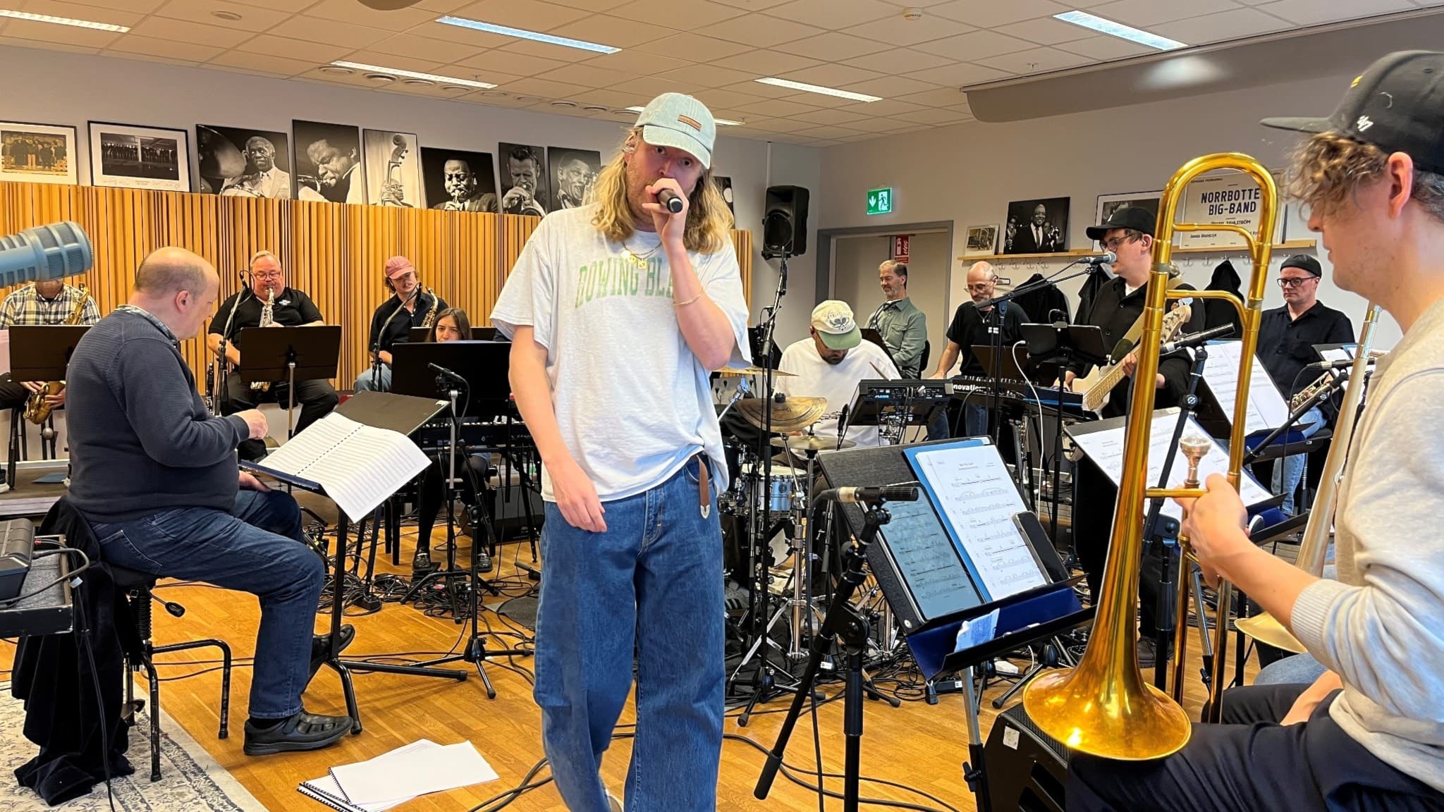 Johan Rensfeldt från Movits! sjunger i en repetitionslokal med Norrbotten Big Band. Han står i centrum medan musiker spelar olika instrument, inklusive trombon och piano, i bakgrunden.