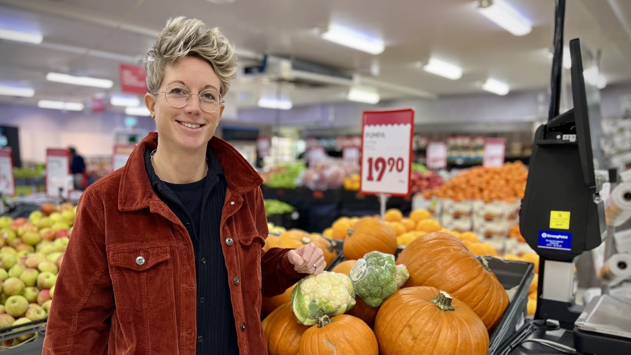 Malin Sand, dietist och folkhälsostrateg på Region Norrbotten, står vid en frukt- och grönsaksdisk i en livsmedelsbutik med pumpor och andra grönsaker i fokus.