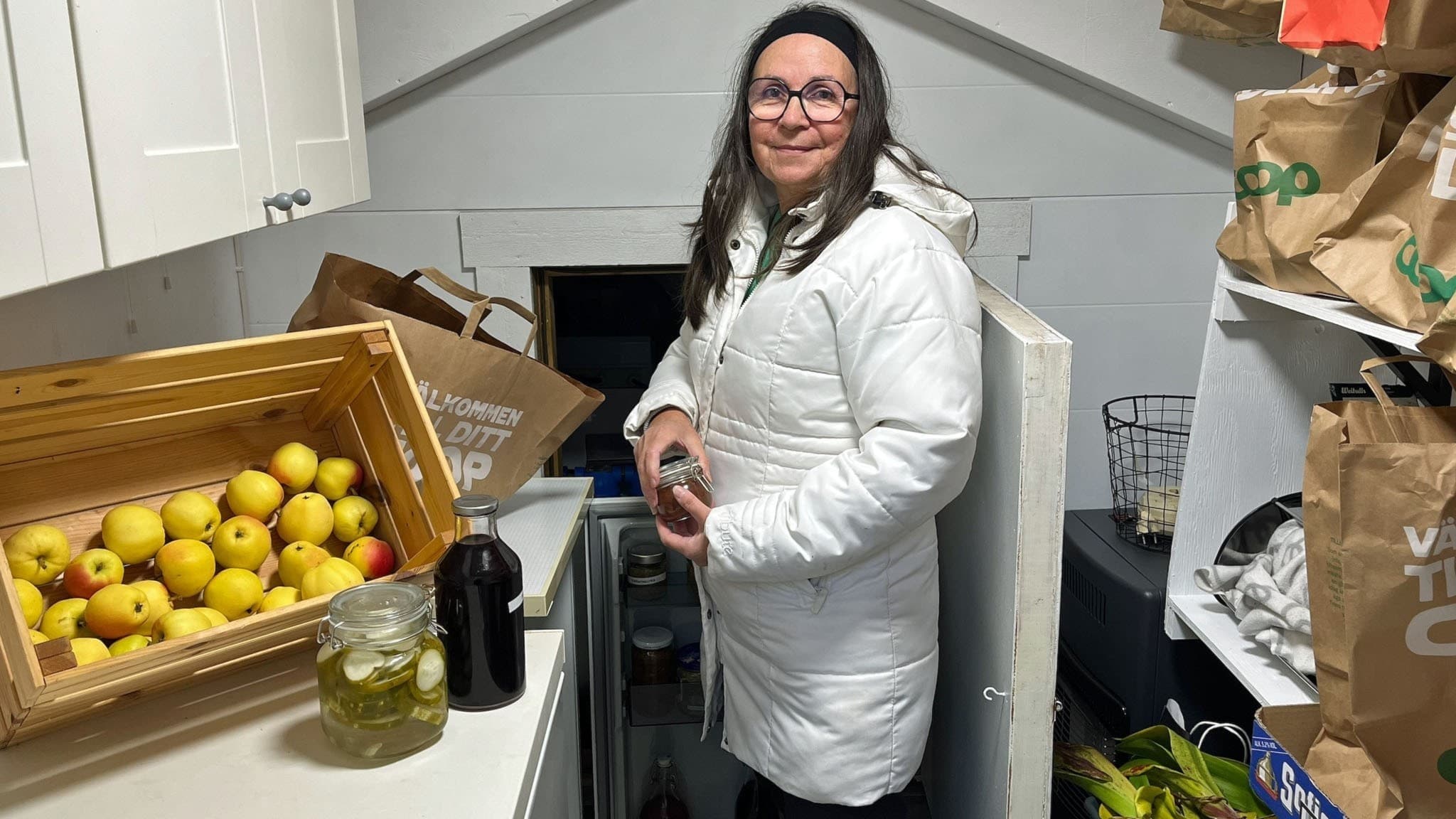 Carina står i sitt förråd, hon har mörkt långt hår och mörka glasögon. I rörrådet syns äpplen i en låda och en massa burkar med  konserverad mat. 