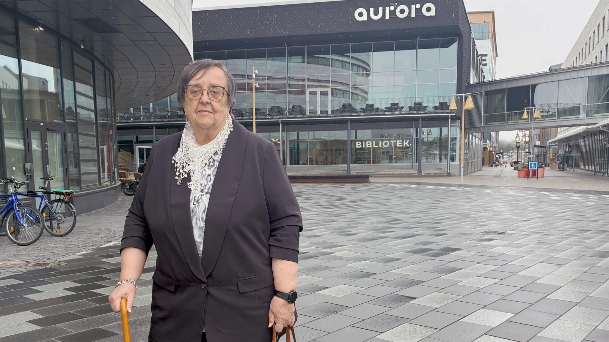 En kvinna står framför Aurora Kultur och Kongress i Kiruna. Byggnaden har en modern design med stora glaspartier och synlig skyltning. Platsen är omgiven av ett stenbelagt torg.