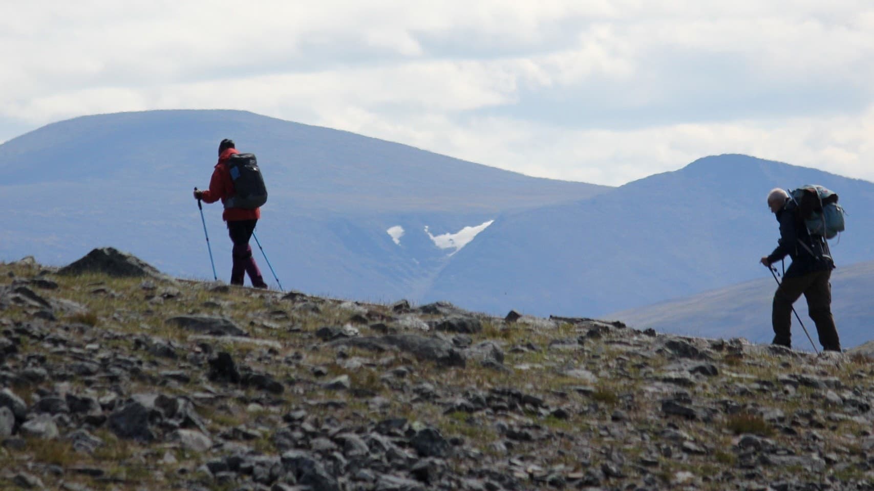 Två fjällvandrare med ryggsäckar och stavar går över en stenig terräng i Tarfaladalen, Kebnekaiseområdet. Bakgrunden domineras av höga berg och en glaciär syns mellan topparna.