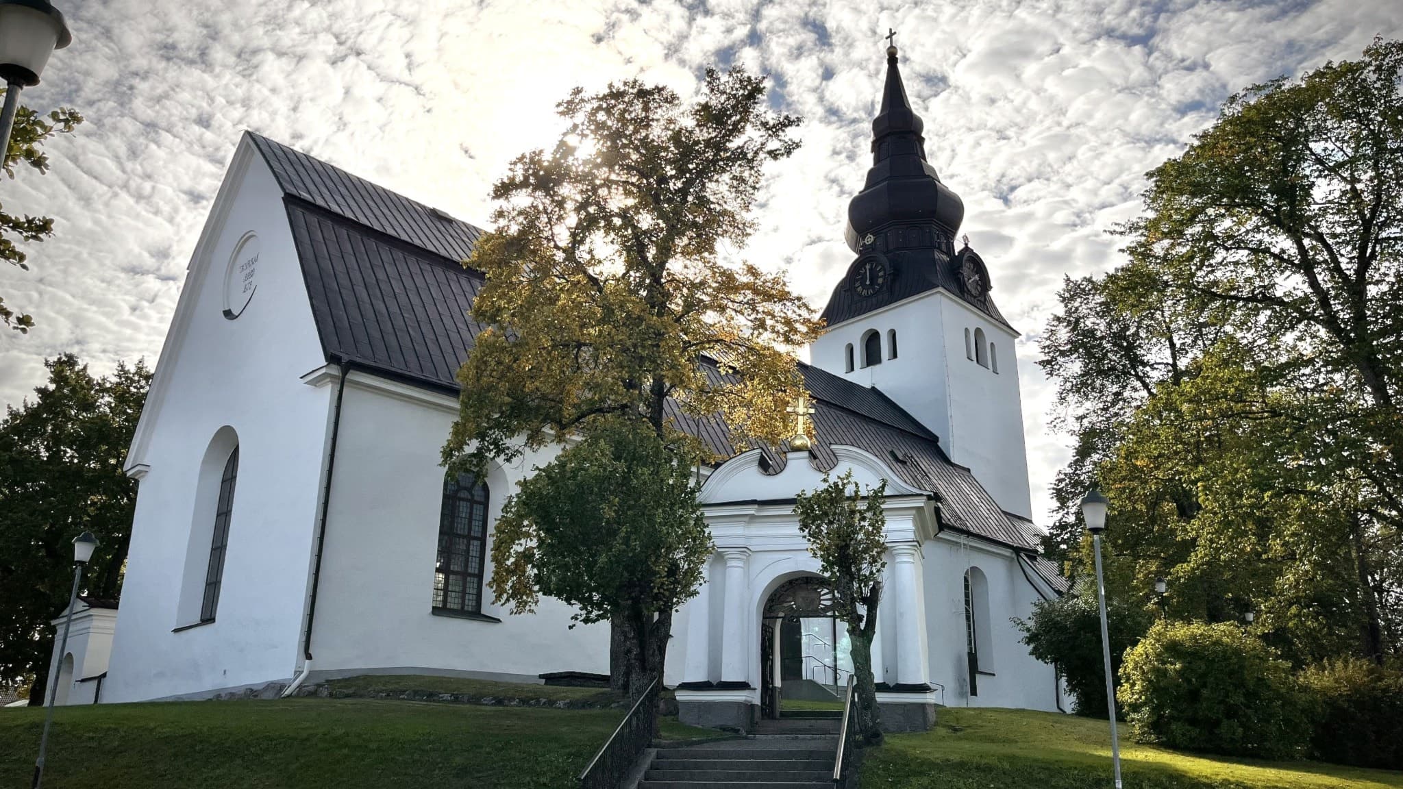En vit kyrka med högt svart torn, omgiven av grönskande träd. Kyrkan är den största i Hudiksvallsbygdens församling, som består av sex kyrkor.