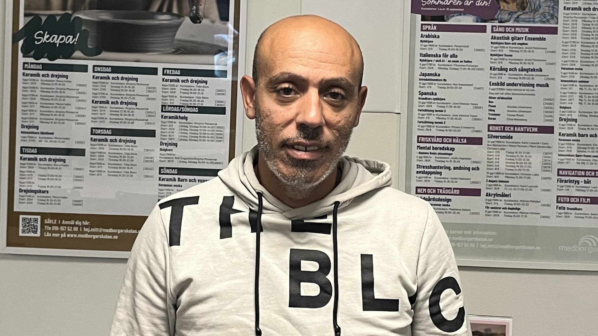 Faris Backar står framför två anslagstavlor med information på. Han har på sig en luvtröja och tittar in i kameran. 
