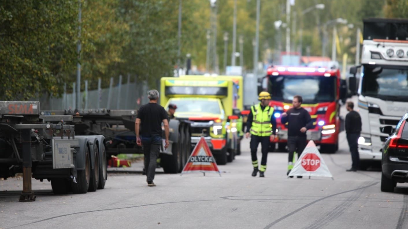 En olycksplats med en motorcykel på en väg. Räddningspersonal, polis och flera utryckningsfordon är på plats. Trafiken är avspärrad med varningsskyltar. 