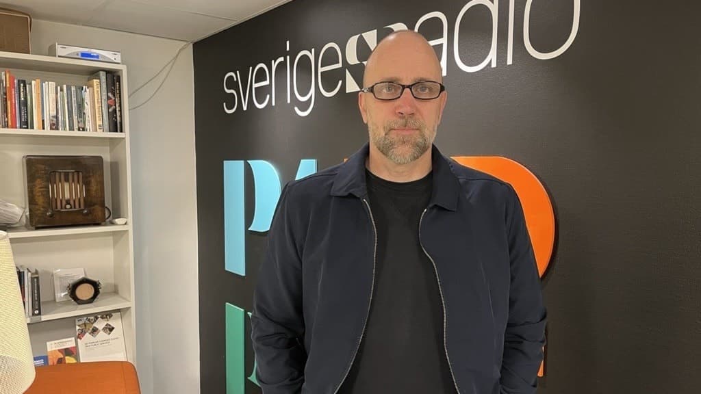 Kommunpolisen Måns Jennehag står framför en vägg med Sveriges Radios logotyp. Han bär en mörk jacka och svart tröja. I bakgrunden syns en bokhylla och färgglada stolar.