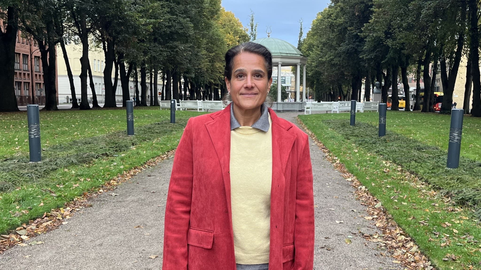 Anna Bakken Krentze står på en grusgång omgiven av gröna träd i en park, iklädd en röd kappa och gul tröja. I bakgrunden syns en historisk byggnad med kolonner.