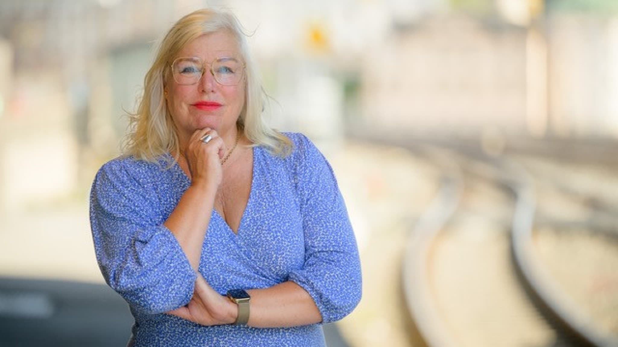 Ingela Bendrot i blå klänning står med handen vid hakan. Till höger syns en järnväg med dubbelspår som sträcker sig genom ett öppet landskap under molnig himmel.