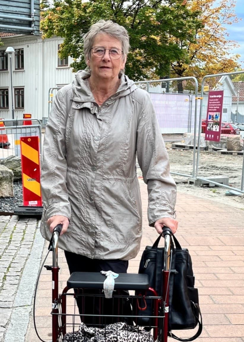Britt Inger Eriksson går med en rullator på en trottoar intill ett pågående vägarbete med avspärrningar och byggstaket i bakgrunden.