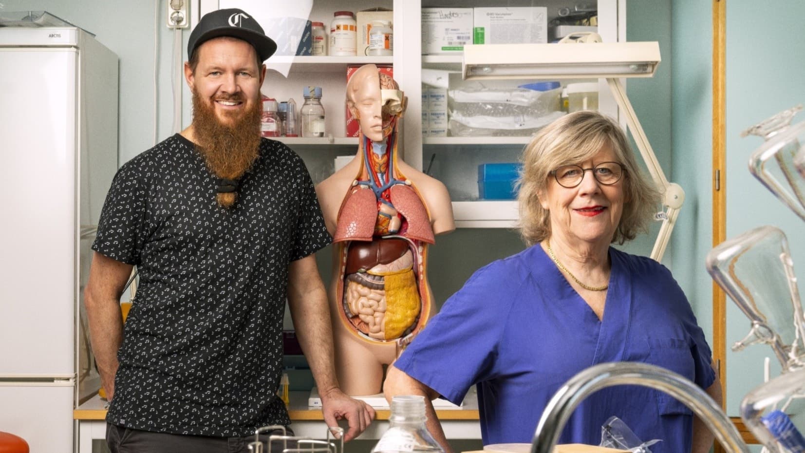 Christer Lundberg och Agnes Wold ståendes i ett laboratorium. Christer med skägg och keps till vänster i bild, till höger om honom en anatomisk modell över en människokropp. I förgrunden, till höger, står Agnes Wold med en blå läkarskjorta och glasögon.