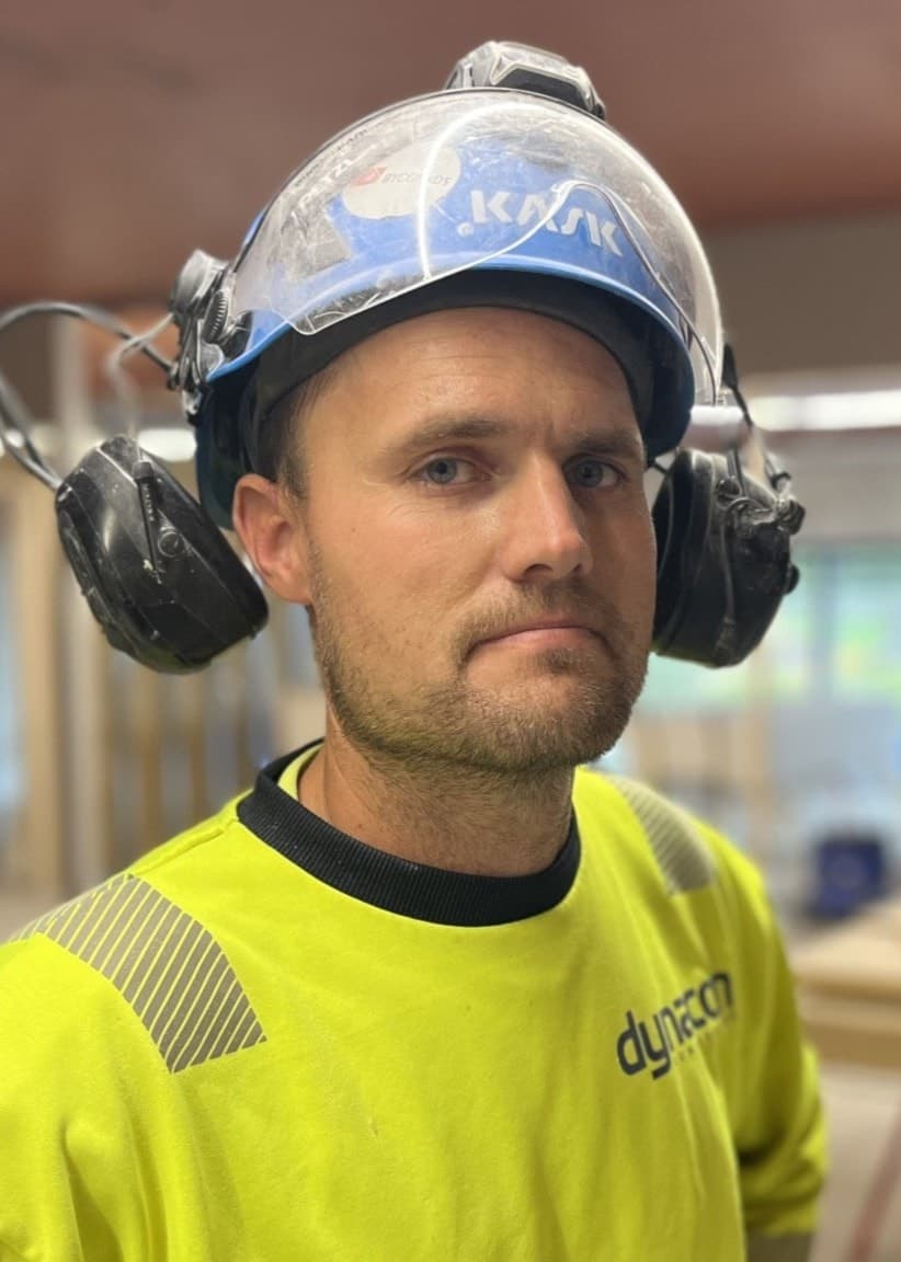 En byggnadsarbetare i gul varseltröja och skyddsutrustning står i ett pågående byggprojekt med träbalkar och verktyg synliga i bakgrunden. Snickaren Michael vill också ha kortare arbetsdag